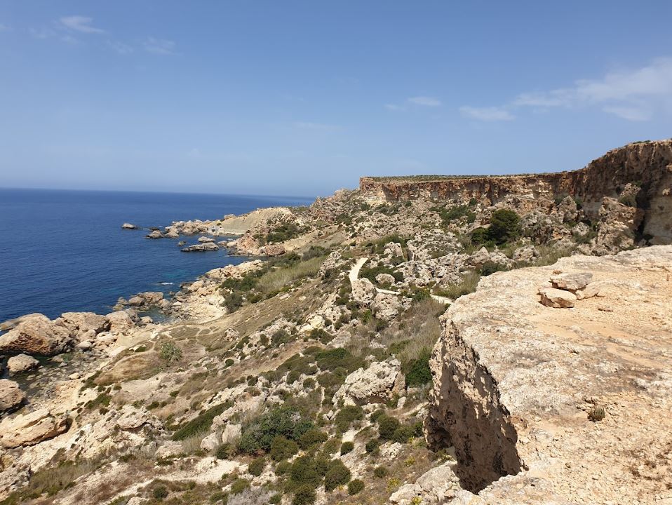 a sandy and rocky coastal landscape in Malta, with the sea on the right and bright blue sky