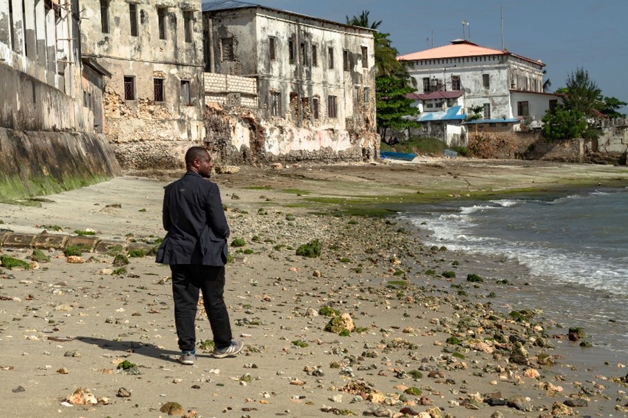 Cydnabyddiaeth am y llun: Makame Juma, Prif Swyddog Gweithredol Cymdeithas Treftadaeth Stone Town Zanzibar, ar lan y môr yn Stone Town. Mae effeithiau erydu a chyrydu gan halen i’w gweld yn glir ar wynebau’r adeiladau cyfagos. 