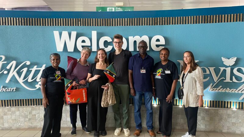 a group of people standing in front of a sign saying welcome to st kitts and nevis, smiling at the camera