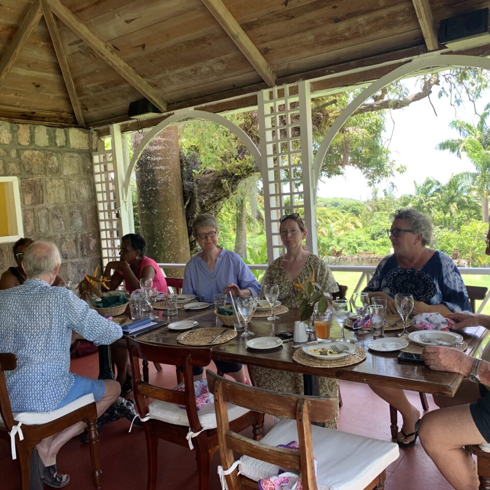 A small group of people having a meeting around a wooden table in St Kitts with a lush green background beyond the veranda behind them