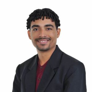 a photo of a man with dark skin and black hair styled in locs, wearing a dark jacket, smiling at the camera