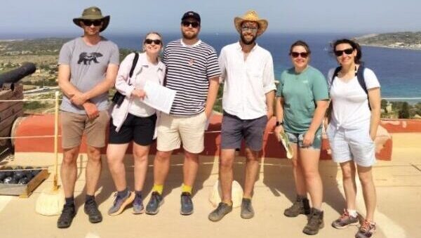 A group of people smiling at the camera, with the backdrop of blue sea and bright blue sky, taken in Malta