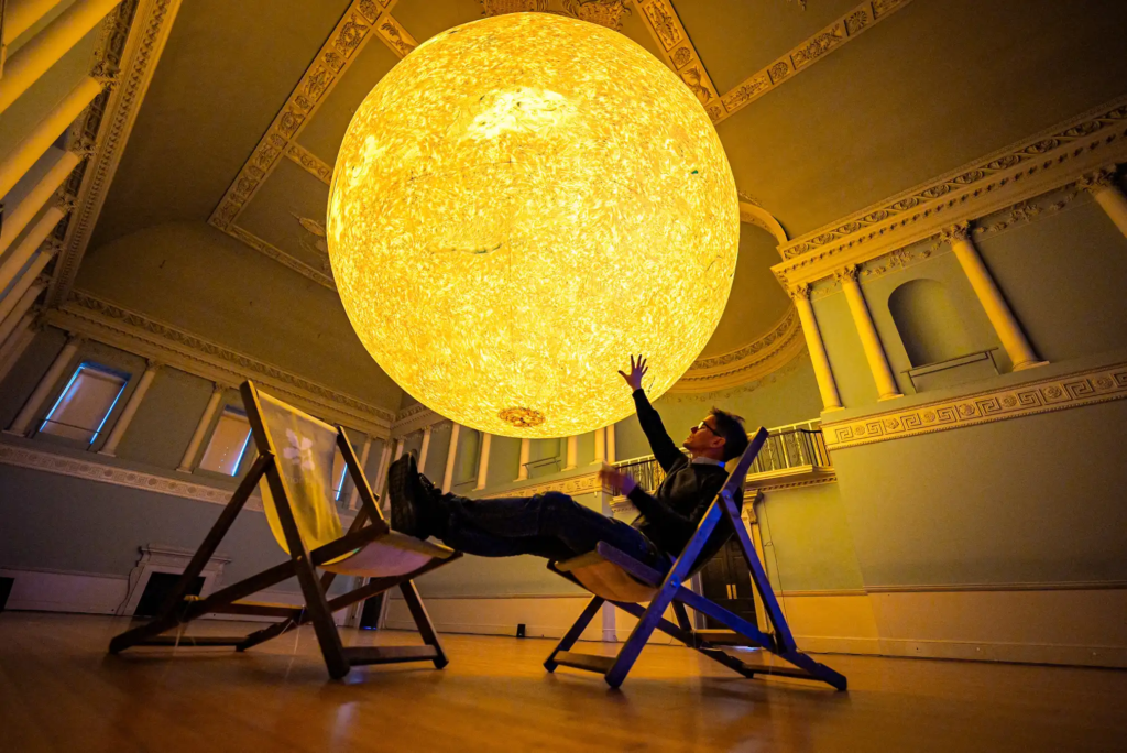 the interior of an historic building with a very large spherical sculpture of the sun hanging from the ceiling. Under the sculpture is the silhouette of a man in a deckchair