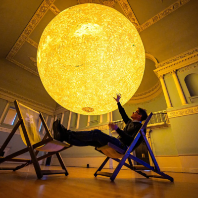 the interior of an historic building with a very large spherical sculpture of the sun hanging from the ceiling. Under the sculpture is the silhouette of a man in a deckchair