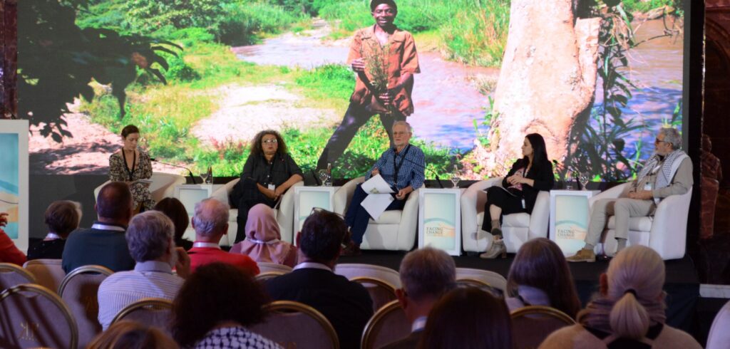 a panel of four speakers seated on stage in front of a presentation on a large video screen