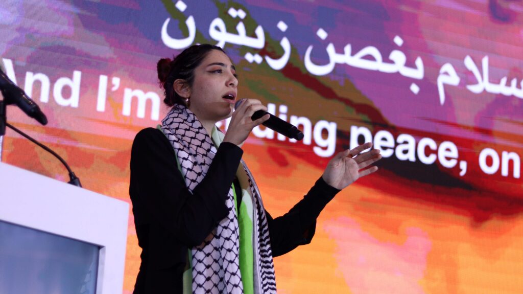 a young woman singing into a microphone. she is standing in front of an orange and purple screen with arabic and english script and wearing a Palestinian keffiyeh scarf