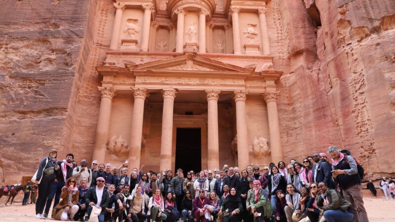 a large group of people smiling at the camera in front of The Treasury, a large temple carved from red stone in Petra, Jordan