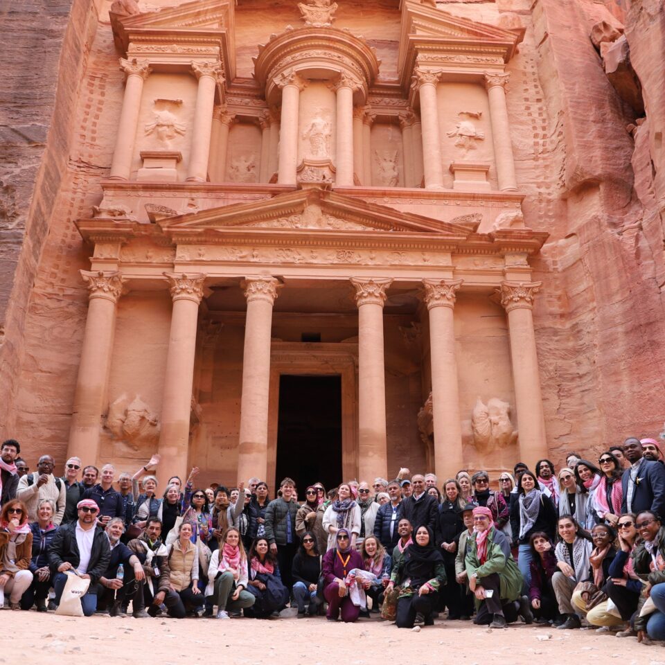 a large group of people smiling at the camera in front of The Treasury, a large temple carved from red stone in Petra, Jordan