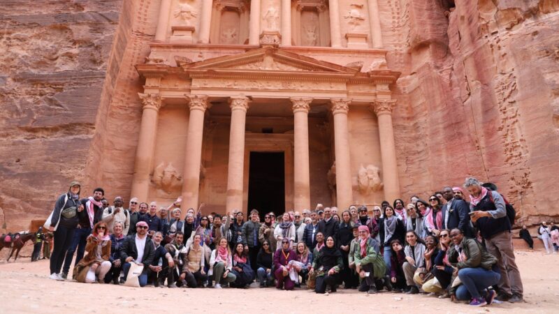 a large group of people smiling at the camera in front of The Treasury, a large temple carved from red stone in Petra, Jordan