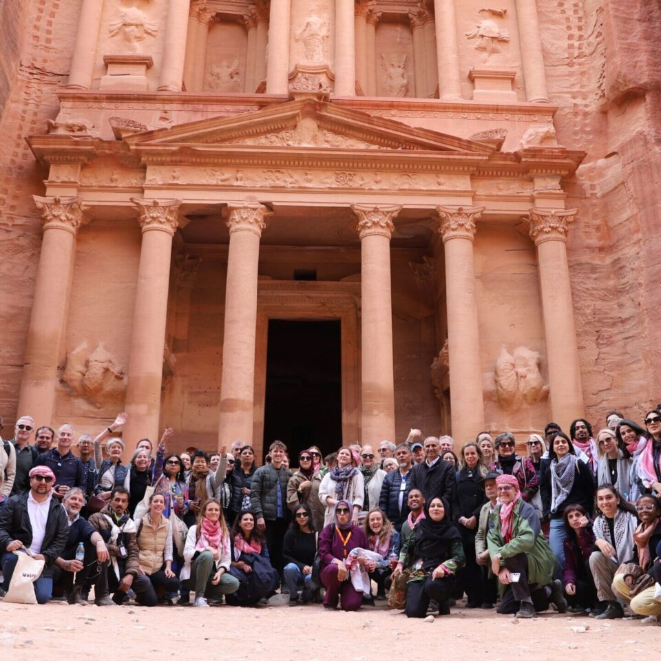 a large group of people smiling at the camera in front of The Treasury, a large temple carved from red stone in Petra, Jordan