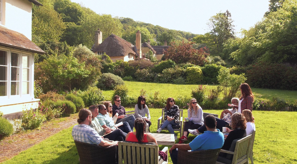 A large group of people sitting in a circle outside a cottage in the sunshine